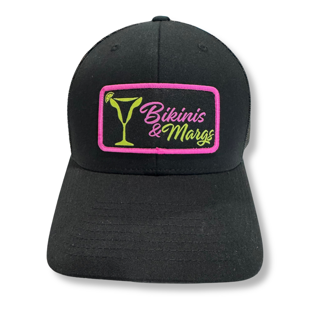 A black baseball cap with a rectangular embroidered patch on the front. The patch features a bright pink border, a green martini glass graphic, and the words 'Bikinis & Margs' in pink and green script.