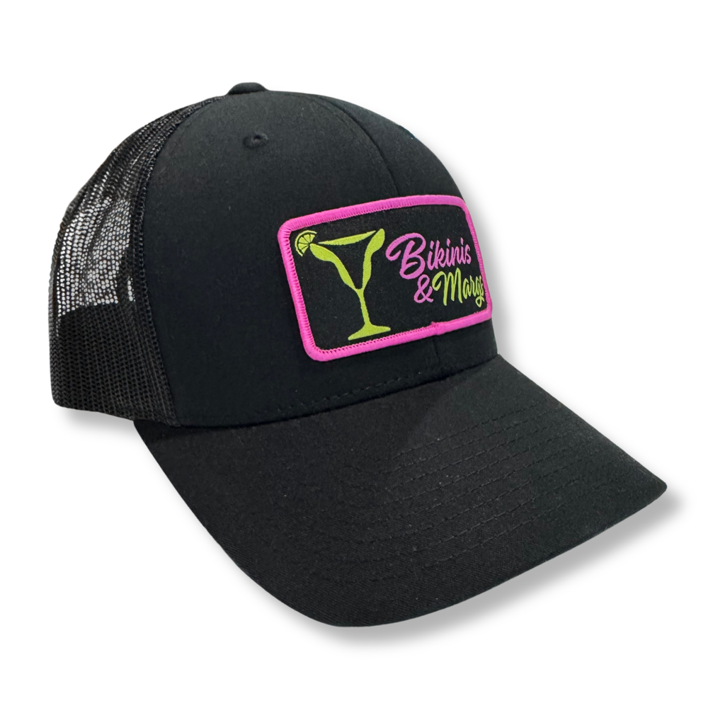 A black trucker hat with a curved brim and black mesh back panels, featuring a rectangular embroidered patch on the front with a pink border, a green martini glass, and the text 'Bikinis & Margs' in pink and green.