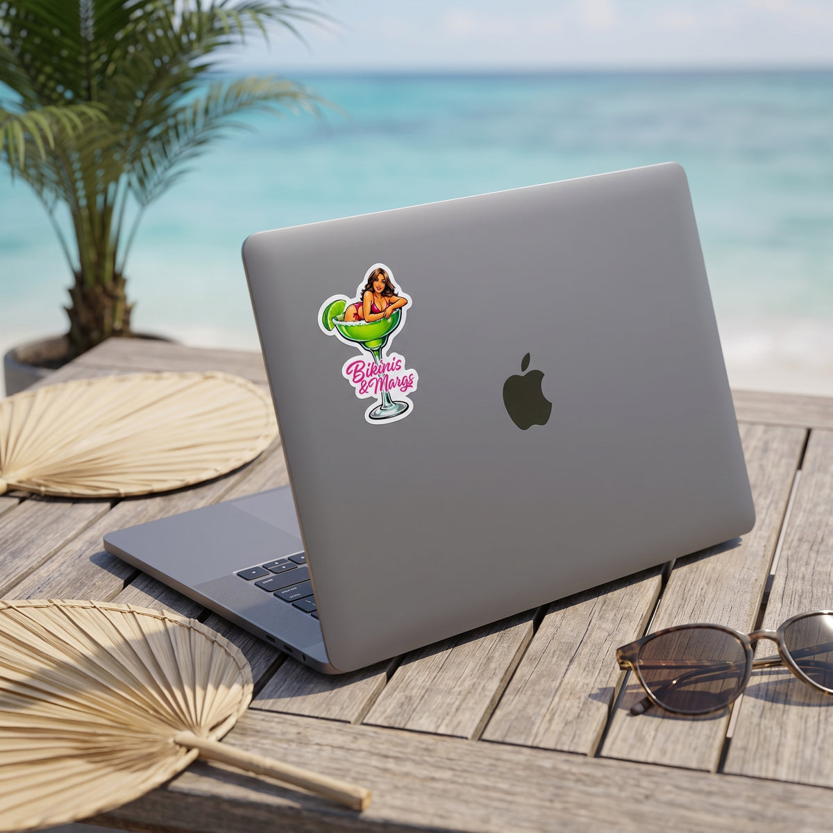 A silver laptop with a colorful 'Baja Vibes' sticker on the back is placed on a wooden table with a tropical beach background. The laptop is styled with a palm fan, sunglasses, and a potted palm. Featuring a sticker with a pin-up style woman sitting in a margarita glass with the words Bikinis & Margs in pink.