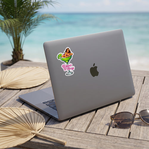 A silver laptop with a colorful 'Baja Vibes' sticker on the back is placed on a wooden table with a tropical beach background. The laptop is styled with a palm fan, sunglasses, and a potted palm. Featuring a sticker with a pin-up style woman sitting in a margarita glass with the words Bikinis & Margs in pink.