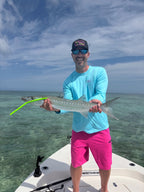 A man wearing a light blue long-sleeve shirt, pink shorts, and a black Bikinis & Margs hat stands on a boat holding a large fish over clear turquoise water under a blue sky.