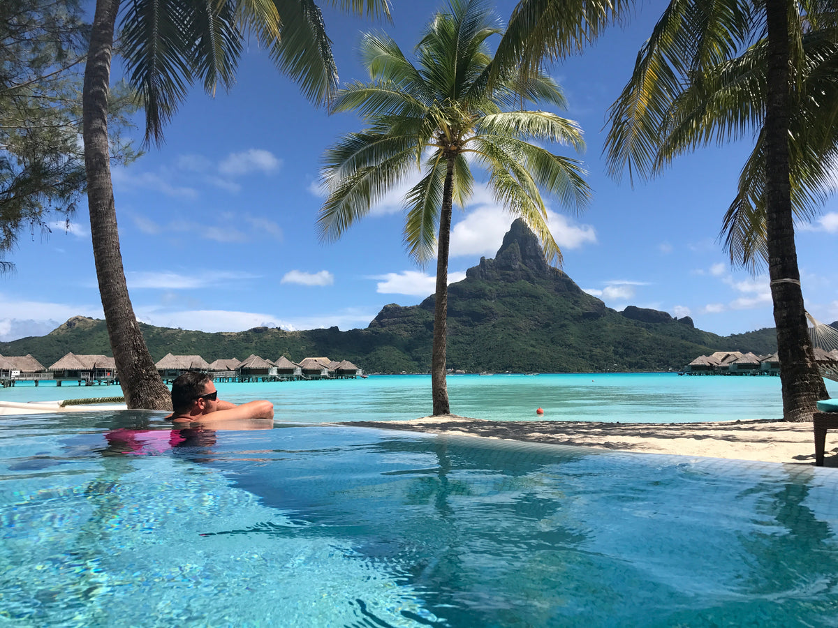 Person relaxing in a pool with palm trees and over the water bungalows and mountain in the background