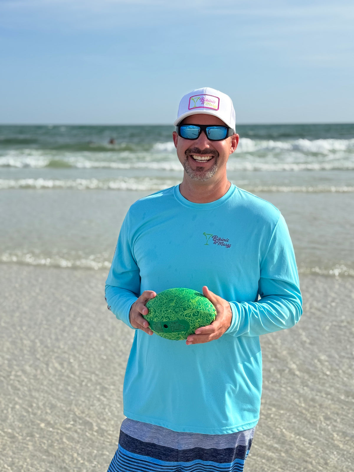 A man standing on a sandy beach wearing a light blue long-sleeve shirt, a white Bikinis & Margs hat, and sunglasses while holding a green textured ball. The ocean and blue sky are visible in the background.