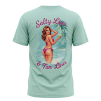 A mint green short-sleeve t-shirt shown from the back, featuring a large graphic print of a retro pin-up woman in a pink bikini on a tropical beach with the text 'Salty Lips & Tan Lines' in pink script.